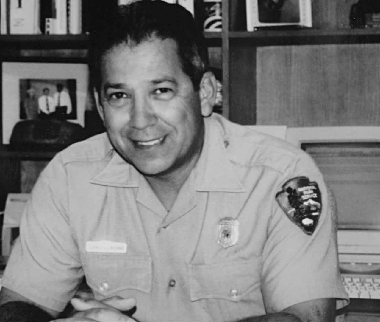 Black and white photograph of Ernest Quintana in National Park Service uniform indoors, seated at desk