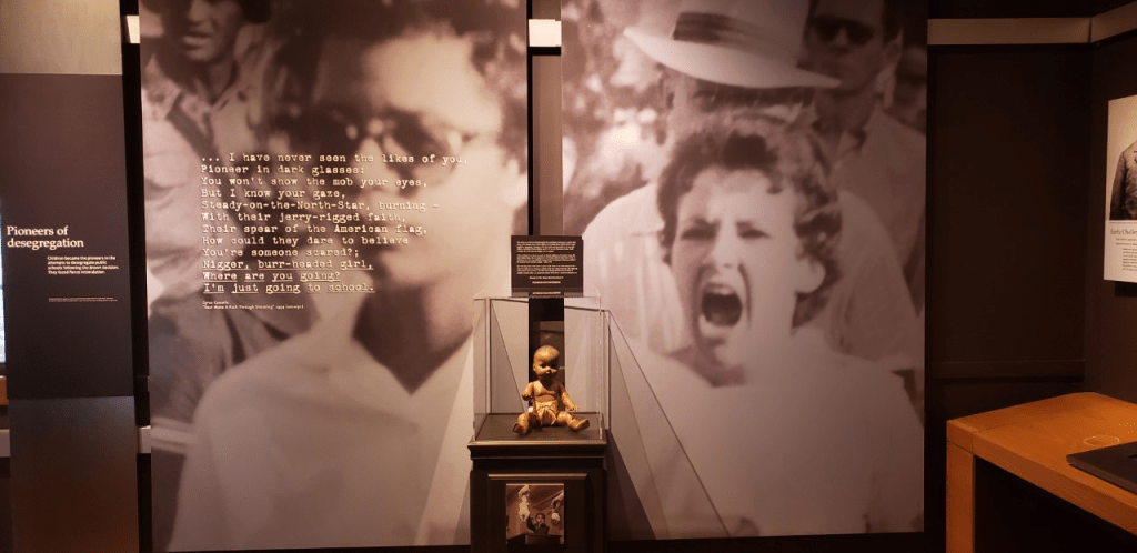 One of four dolls used in the experiment conducted by psychologists Kenneth and Mamie Clark that would later be used in Brown v. Board of Education on display in a glass case in front a photo of Elizabeth Eckford, being verbally attacked by an angry mob, outside of Little Rock Central High School in 1957.