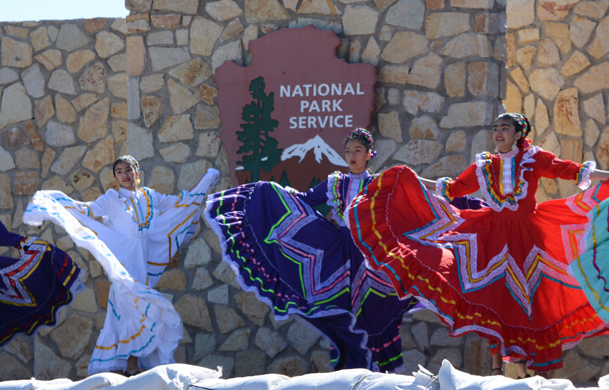 Dancers wearing colorful, traditional Mexican dresses perform on a stage in front of a National Park Service arrowhead.